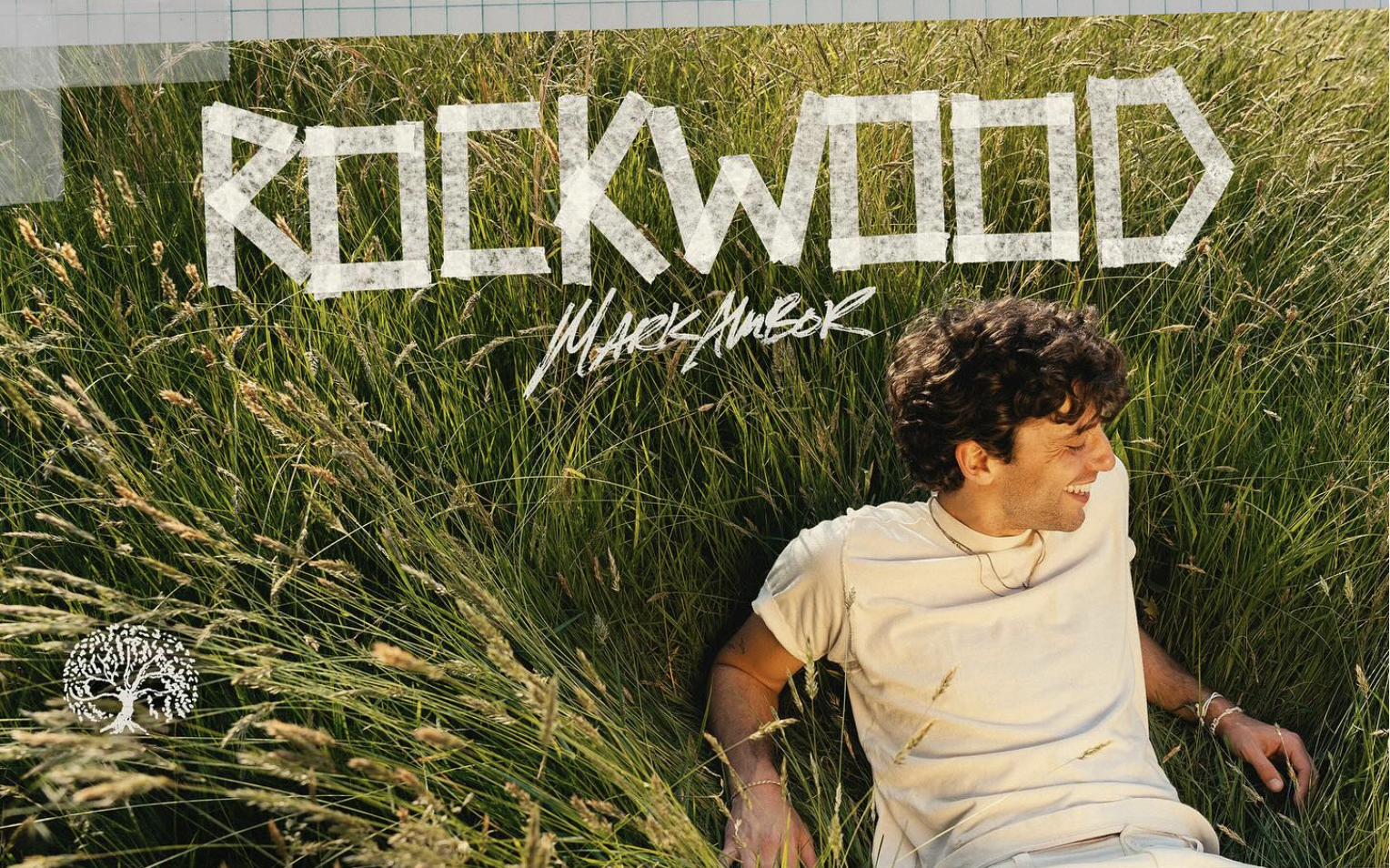 Mark laying on grass, wearing a white t-shirt. Text above says 'Rockwood' in all caps and his name in cursive blow.